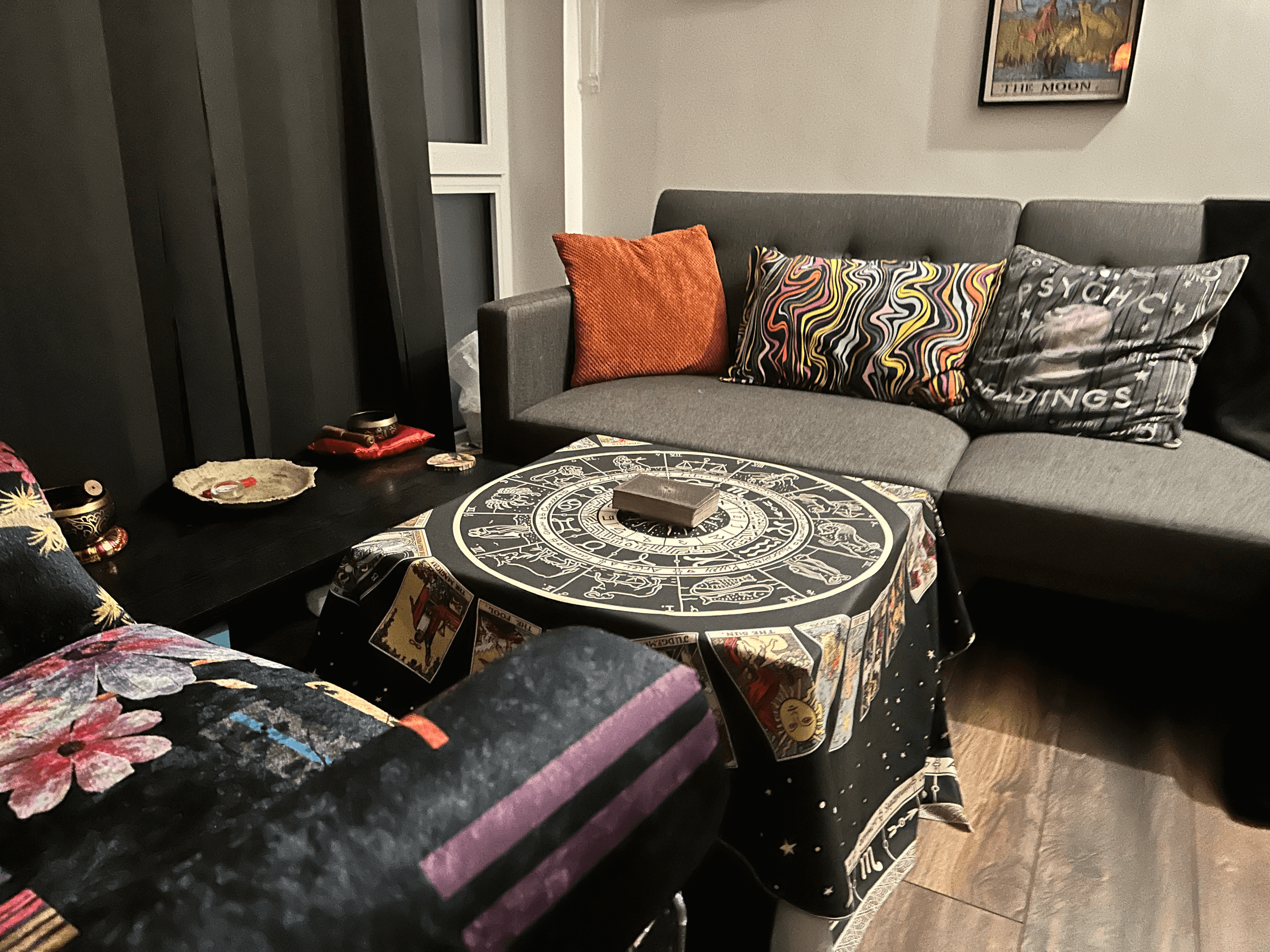 This image gives a closer look at the cosy tarot reading setup within the tarot with gord sanctuary. The focal point is the round table, beautifully draped with a cloth featuring astrological and tarot imagery, with a deck of cards waiting for the next reading. The grey sofa in the background, accented with vibrant cushions—one of which reads “psychic readings”—adds a touch of personality to the room. The plush chair nearby features colourful floral designs, offering an additional comfortable seat. On the side table, small ritual items like a bowl and incense burners sit quietly, ready to enhance the spiritual experience. With tarot-themed artwork on the walls and soft lighting, the space feels both grounded and mystical—ideal for reflection, guidance, or spiritual connection.