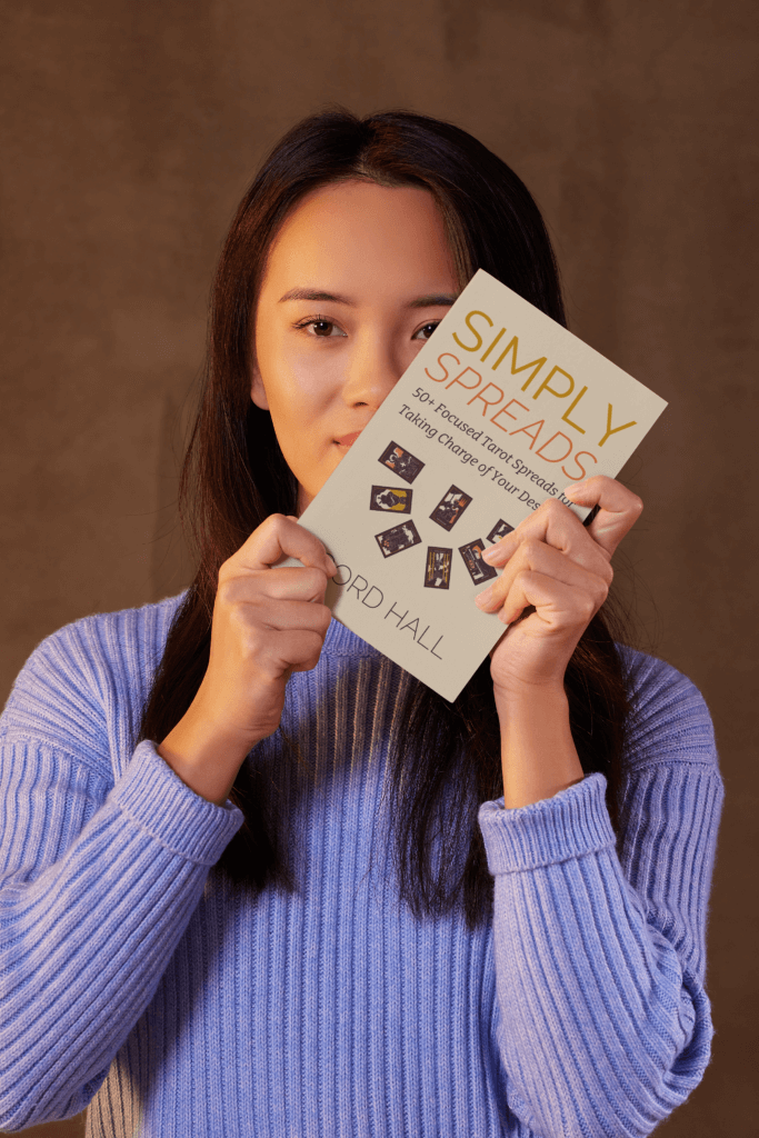 A woman in a blue sweater holding up the book “simply spreads” by gord hall in front of her face, showcasing its cover. The book features tarot cards on the cover and highlights its focus on tarot spreads for self-empowerment.