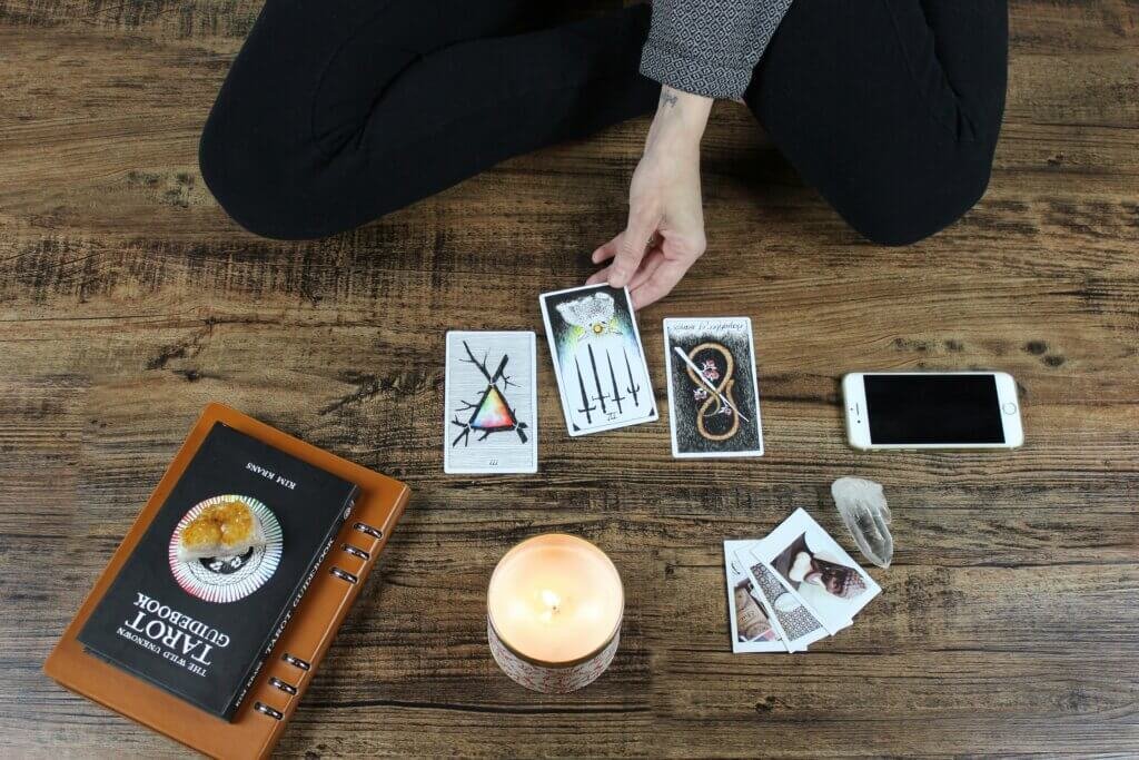 A person performing a tarot reading with three cards laid out on the floor, surrounded by a tarot guidebook, a candle, and crystals. The person’s hand gently touches one of the cards. The setting reflects a grounded, intentional connection to spiritual flow.