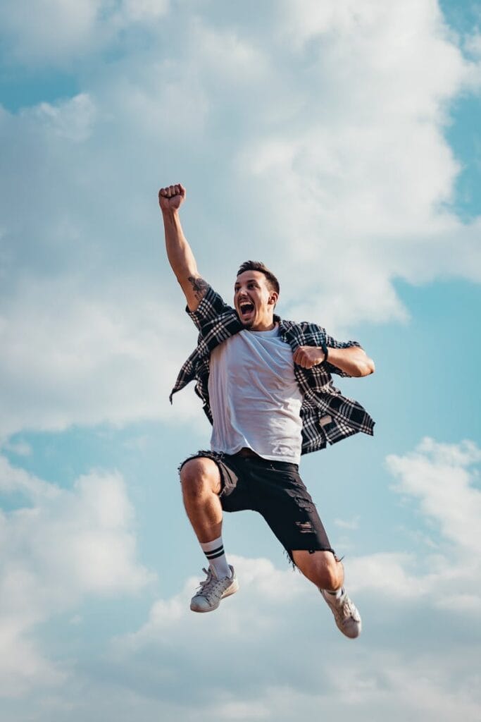 A joyful young man jumps midair with clouds and blue sky in the background, exuding energy and freedom.