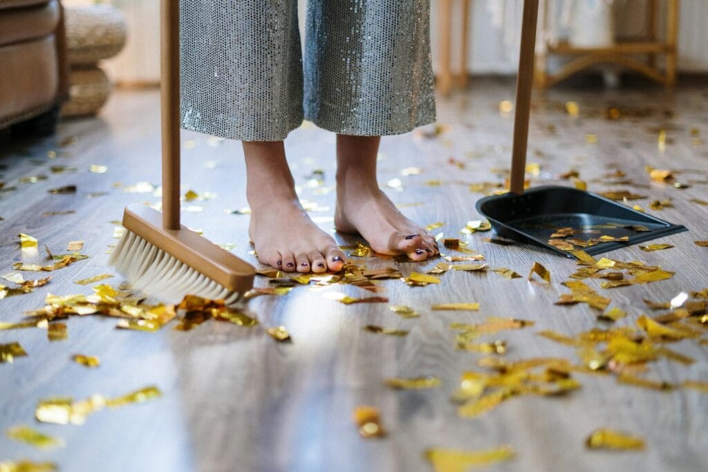 Barefoot woman sweeping gold confetti with broom and dustpan indoors.