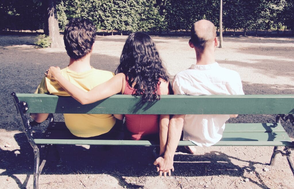 Three people sitting on a park bench. The person in the middle has their arms around both individuals on either side, symbolizing a polyamorous relationship. One person on the end is holding hands secretly with the person in the middle, highlighting the complexity of relationships.